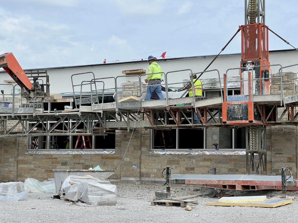 outside incomplete cafeteria. most of the exterior is covered in Lannon stone. Construction workers are standing on scaffolding. April 28, 2025.