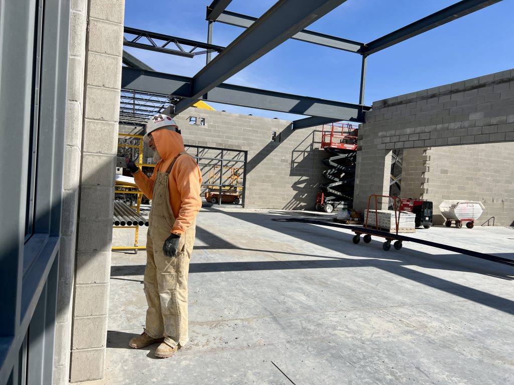 construction worker in orange sweatshirt and overalls working inside incomplete structure. no roof. walls partially complete. April 4, 2025.