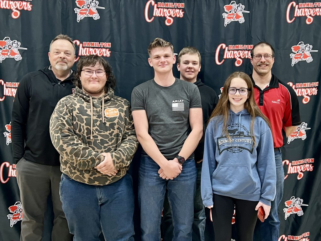 Two teachers and four high school students stand in front of a school banner.